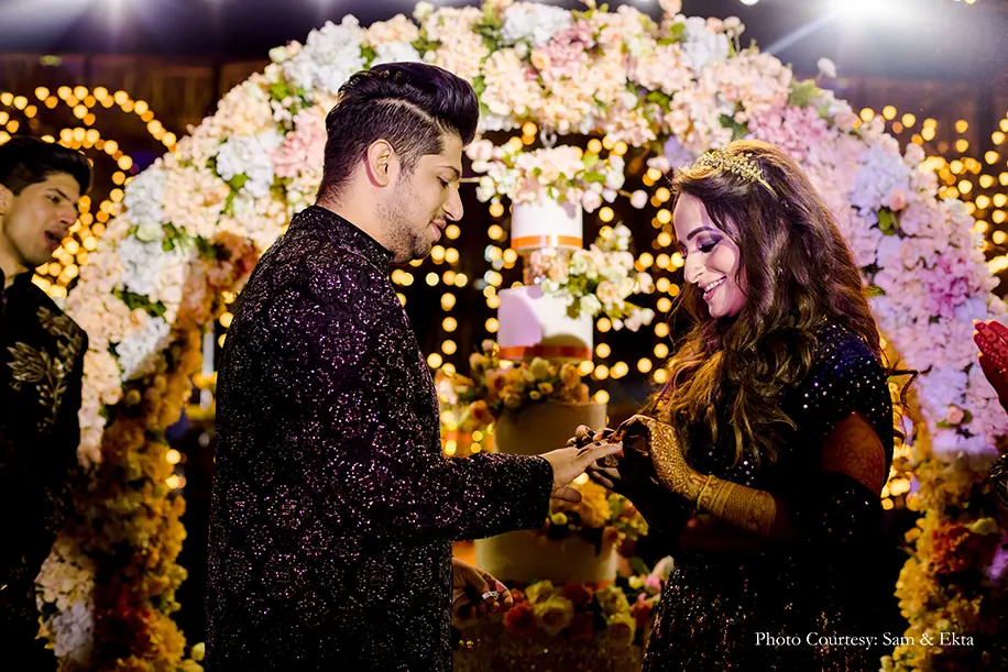 Bride wearing peacock-hued lehenga and groom wearing black and gold sherwani for the sangeet and ring ceremony