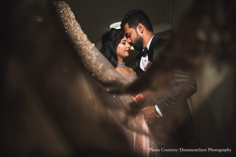 Bride in off-shoulder gown with a huge trail from Kalki and groom in black tuxedo by Nicky Prithyani