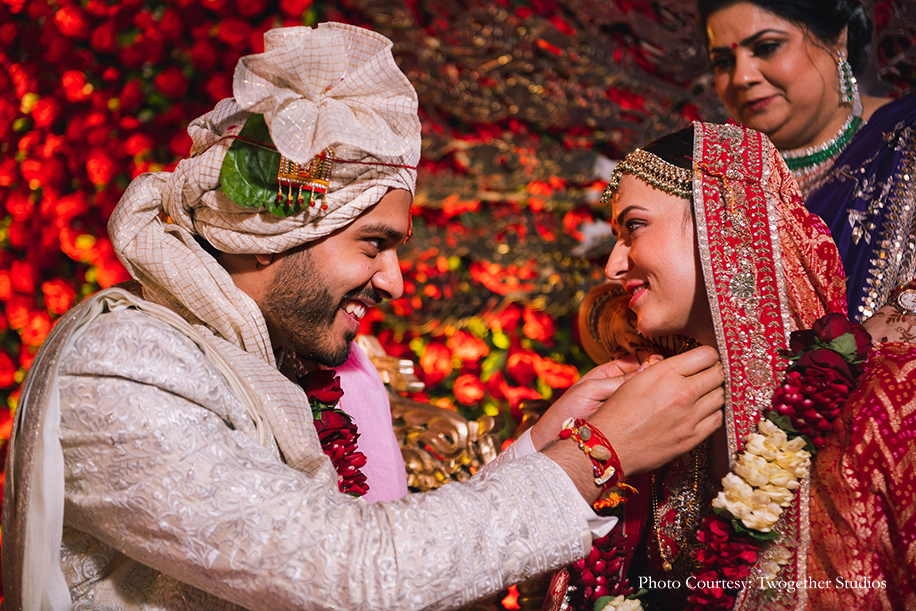 Bride wearing red and gold embroidered lehenga, statement nath and maang tikka for her wedding and Groom wearing white bandhgala fro the wedding at The Leela Ambience, Gurugram