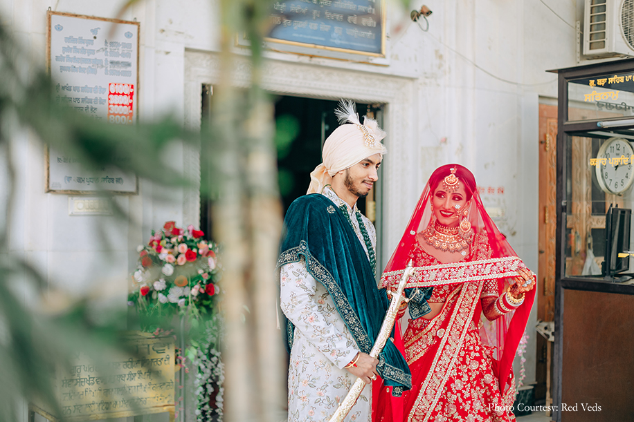 Bride in red lehenga with elaborate polki jewelry and Groom in a white floral sherwani with an emerald green shawl and a multilayered necklace