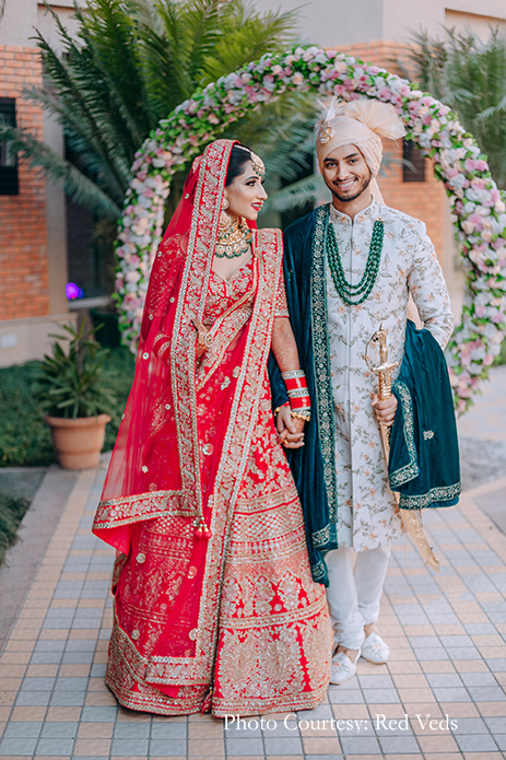 Bride in red lehenga with elaborate polki jewelry and Groom in a white floral sherwani with an emerald green shawl and a multilayered necklace