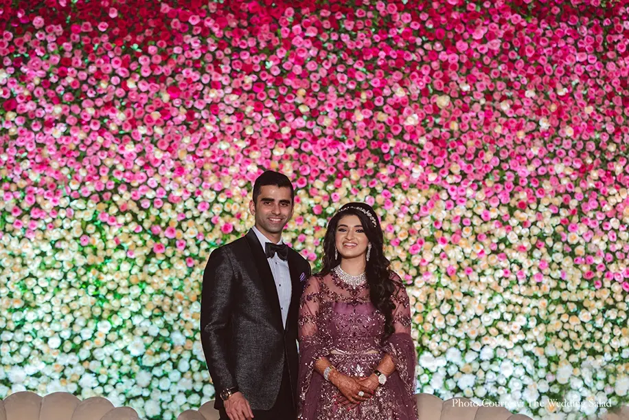 Bride wearing wine gown and groom wearing black tuxedo for the reception posing against a gigantic ombre wall of pink blooms